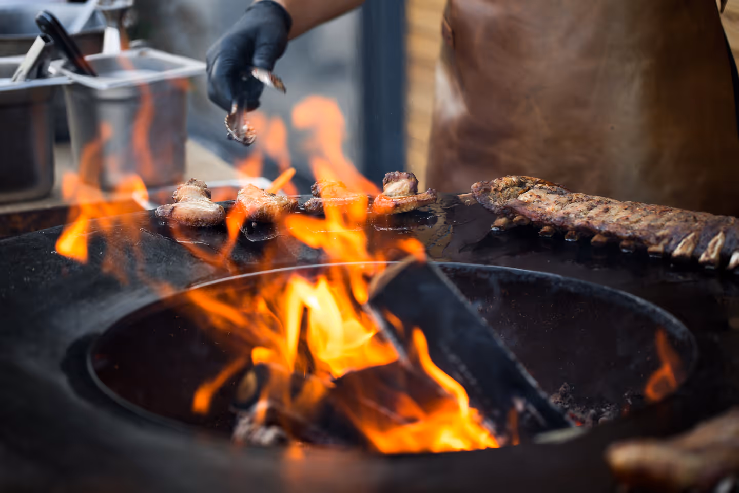 Grilled pork baby ribs and chicken wings cooking over an open flame with a person wearing a leather apron and black glove tending the grill.